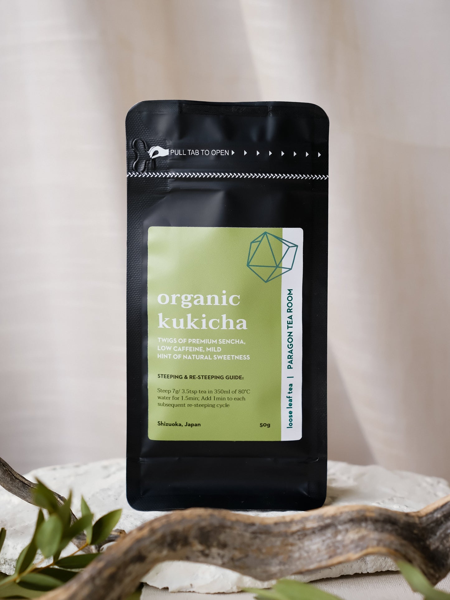 Black package of organic kukicha tea on a neutral background with green leaves.